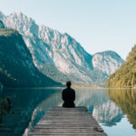 A man sitting on a dock in front of mountains. by Chasing the Sun Vacations