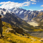 A man standing on the side of a mountain overlooking a lake, contemplating his upcoming cruise adventure. by Chasing the Sun Vacations