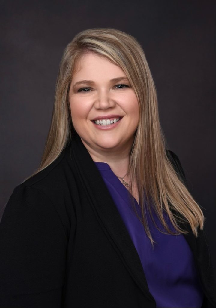 Professional portrait of a smiling woman with long blonde hair wearing a black blazer and purple blouse. by Chasing the Sun Vacations