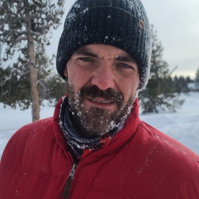 A man wearing a red jacket and beanie in the snow. by Chasing the Sun Vacations